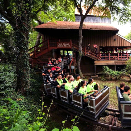 A smiling woman and man enjoy a rollercoaster ride, with others raising their hands in the background.