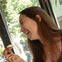 A person smiling while holding a pastry, sitting near a window with trees visible outside on a sunny day.