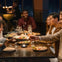 A group of people sit around a dining table, sharing a meal and smiling, with a night cityscape visible through the windows.