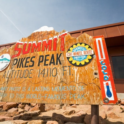 A weathered wooden sign at Pikes Peak Summit showing altitude, surrounded by rocks and a building in the background.