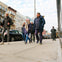 Four people walking on a city sidewalk, dressed in winter clothing, with parked cars and buildings in the background.
