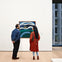 Two people stand in an art gallery, viewing a colorful painting on a white wall. A window shows a cityscape in the background.