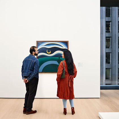 Two people stand in an art gallery, viewing a colorful painting on a white wall. A window shows a cityscape in the background.