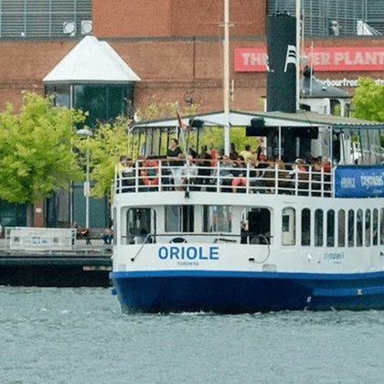 A white ferry named Oriole on the water, with passengers on the upper deck, near a dock with buildings and trees.