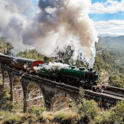 A green steam train emitting white smoke crosses a stone viaduct in a scenic, hilly landscape under a partly cloudy sky.