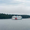 A ferry with 'BC Ferries' branding sails on a calm body of water, with a forested shoreline in the background under a cloudy sky.