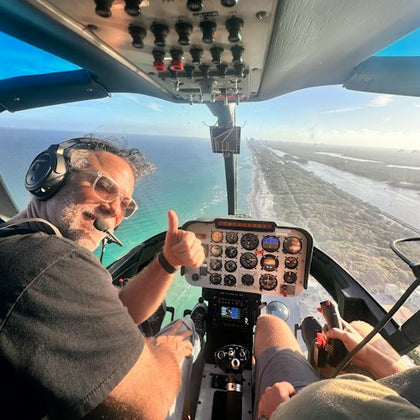 A person wearing a headset gives a thumbs-up inside a helicopter cockpit flying over a coastline.