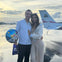 A couple stands on a wet tarmac near a small airplane; the man holds a birthday balloon and gift bag as they pose smiling.