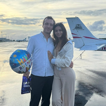 A couple stands on a wet tarmac near a small airplane; the man holds a birthday balloon and gift bag as they pose smiling.