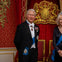 An elderly couple in formal attire stands in front of a red backdrop with a golden crest and two ornate chairs.