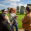 A group of people standing and talking in front of Stonehenge on a sunny day with green grass and a blue sky.