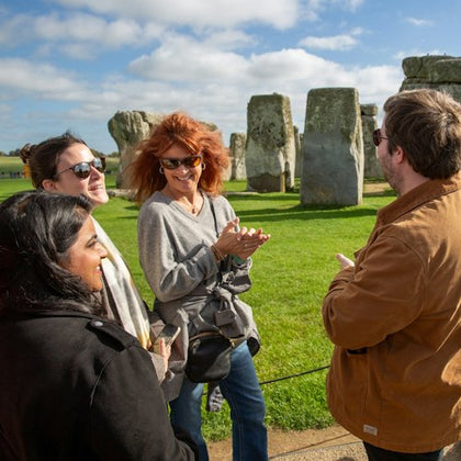 A group of people standing and talking in front of Stonehenge on a sunny day with green grass and a blue sky.