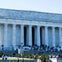 The image shows a large neoclassical building with columns, with many people gathered on its steps and lawn under a clear sky.