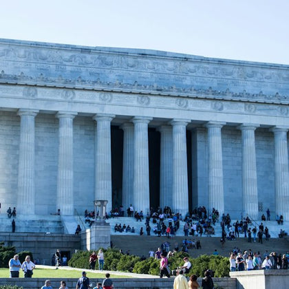 The image shows a large neoclassical building with columns, with many people gathered on its steps and lawn under a clear sky.