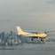 A small airplane flying over a cityscape with tall buildings and a bridge over the water below, under a cloudy sky.