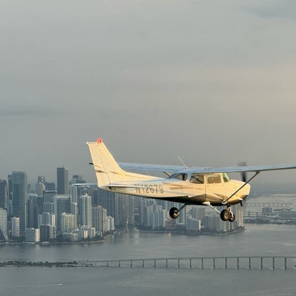 A small airplane flying over a cityscape with tall buildings and a bridge over the water below, under a cloudy sky.