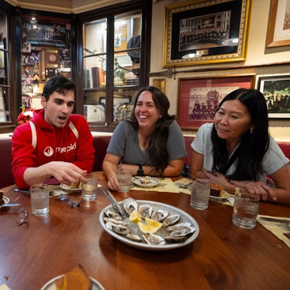 Three people are seated around a table in a restaurant, enjoying a meal of oysters served on a plate with lemon wedges.