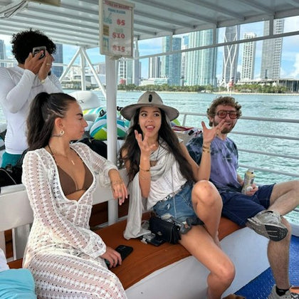 Four people are sitting on a boat with city skyscrapers in the background. One person covers his face while the others are engaged in conversation.