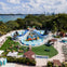 Aerial view of a water park with a large blue inflatable slide, surrounded by trees, picnic tables, and a waterfront cityscape in the background.