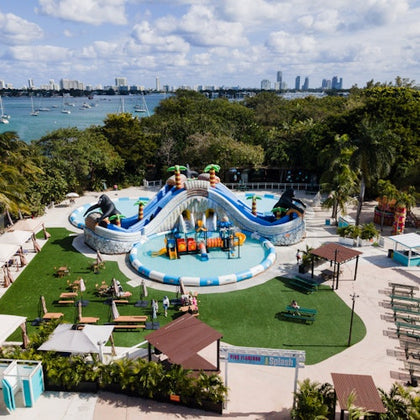 Aerial view of a water park with a large blue inflatable slide, surrounded by trees, picnic tables, and a waterfront cityscape in the background.