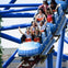 A group of five young people laughing and walking arm in arm at an amusement park with roller coasters and American flags in the background.