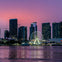 City skyline at dusk with illuminated Ferris wheel and reflections on a calm body of water.