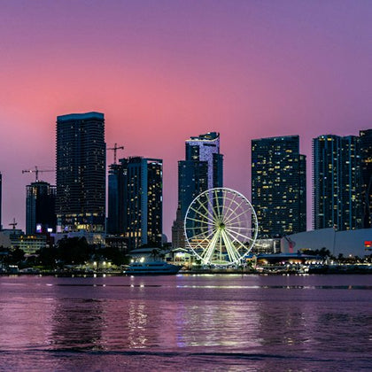 City skyline at dusk with illuminated Ferris wheel and reflections on a calm body of water.