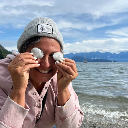 Person wearing a beanie and holding seashells over their eyes, smiling at a beach with mountains and sky in the background.