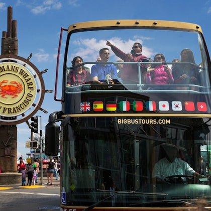 A double-decker tour bus with passengers on the top deck near a Fisherman's Wharf sign in San Francisco.