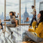 People seated and standing in a modern cafe with large windows showcasing a city skyline, including the Empire State Building.