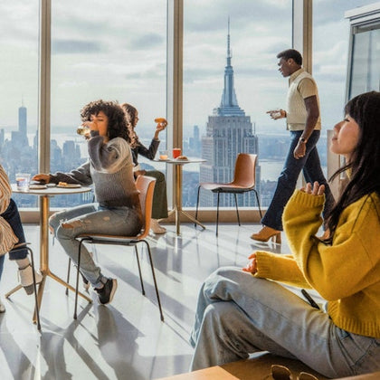 People seated and standing in a modern cafe with large windows showcasing a city skyline, including the Empire State Building.