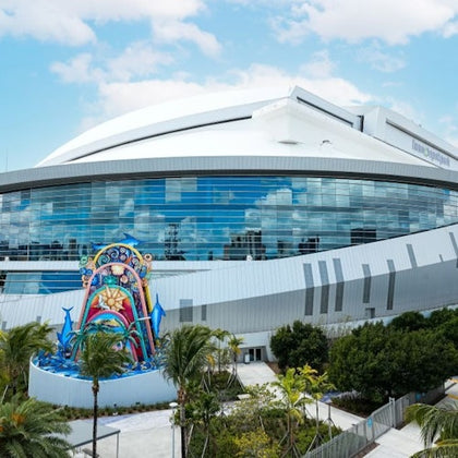 A modern stadium with a curved white roof and glass facade, featuring a colorful sculpture and surrounded by palm trees.