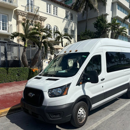 A white van is parked on the street in front of a building with palm trees and balconies under a sunny sky.