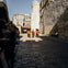 Four guards in red uniforms and one in a traditional outfit march along a cobblestone street, flanked by onlookers.