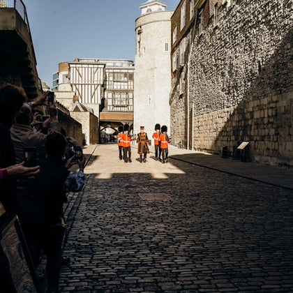 Four guards in red uniforms and one in a traditional outfit march along a cobblestone street, flanked by onlookers.