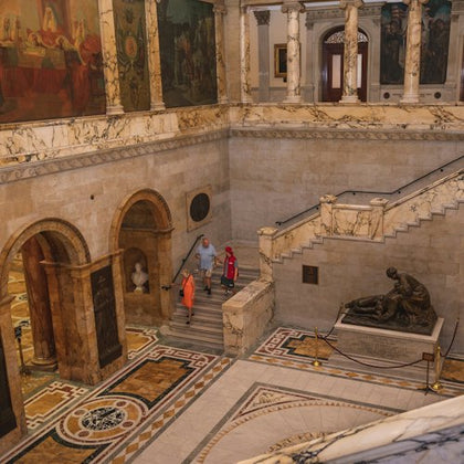 People walking in a marble-floored museum with statues, archways, and large paintings on the walls.