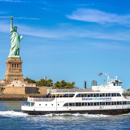 A ferry labeled 'Statue City Cruises' passes in front of the Statue of Liberty on a sunny day.