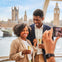A smiling couple holding champagne flutes pose for a photo with the Houses of Parliament and Big Ben in the background.