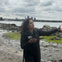 A person standing on a rocky shore under a cloudy sky, gesturing with their hands. A city skyline is visible across the water.