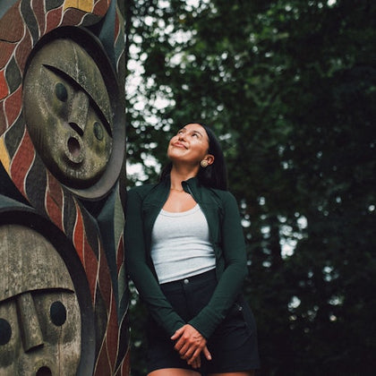 A woman standing next to a carved wooden totem pole, looking upwards. The background features trees and foliage.