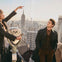 A woman and a man stand on an observation deck overlooking a city skyline, with a viewing telescope in the foreground.