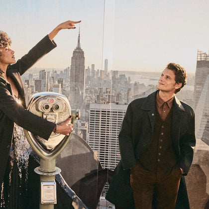 A woman and a man stand on an observation deck overlooking a city skyline, with a viewing telescope in the foreground.