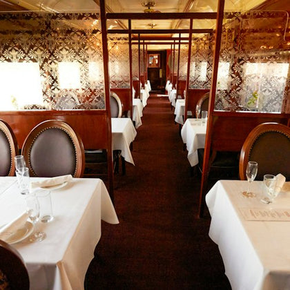 An elegant train dining car with tables set with white linens and chairs, featuring ornate floral-patterned walls.