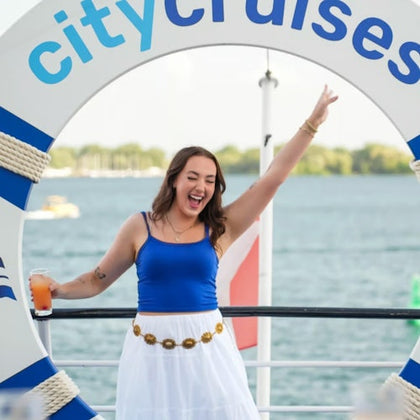 A woman in a blue top and white skirt poses cheerfully with a drink, framed by a life preserver labeled 'City Cruises.'