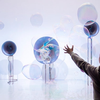 A person reaches towards large iridescent bubbles floating near spherical objects on stands in a bright space.