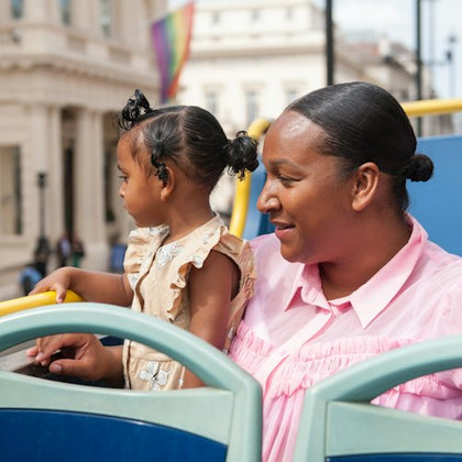 A woman and a young girl sit on an open-top bus, with a cityscape and rainbow flag visible in the background.