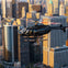 A black helicopter flying over a cityscape with tall buildings and skyscrapers at sunset.