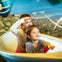 Two smiling children sit in a white boat-shaped structure, pretending to navigate, with a colorful mural in the background.