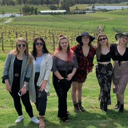 A group of six women posing together on a grassy field with vineyards and a landscape in the background.