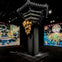 A gallery installation featuring a golden sculpture on a dark pillar and colorful, intricate artworks on the walls.